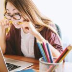 woman biting pencil while sitting on chair in front of computer during daytime